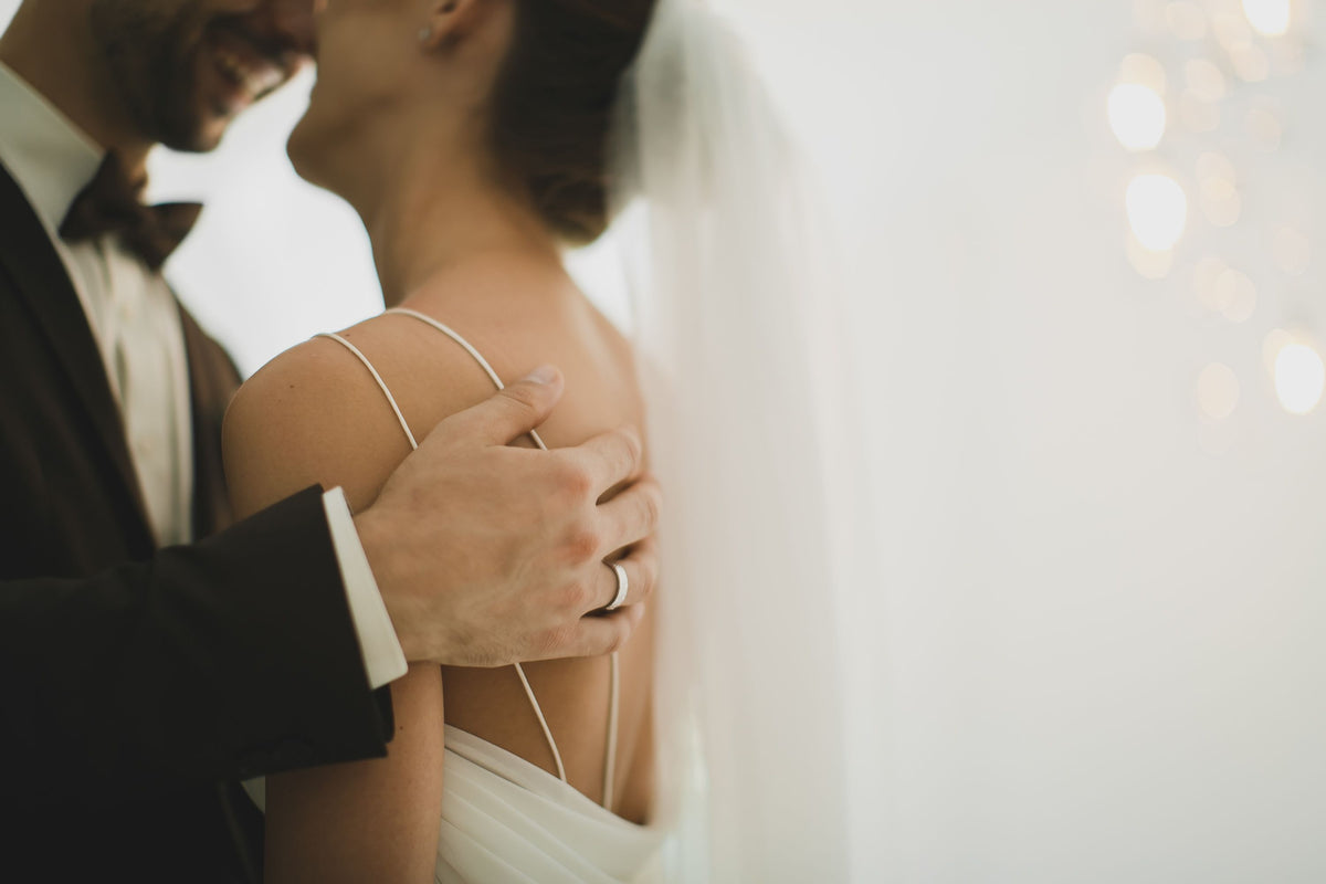 A close-up lifestyle shot of a bride in a white wedding dress with thin straps and a veil, wearing a silver band on her finger, embraced by a groom in a black tuxedo.
