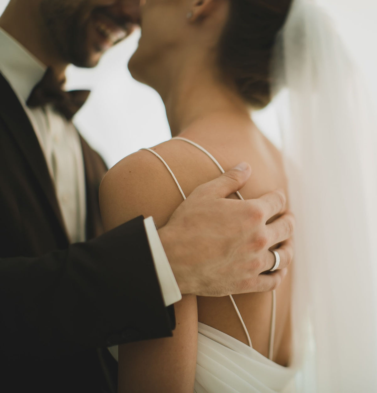 A close-up lifestyle shot of a bride in a white wedding dress with thin straps and a veil, wearing a silver band on her finger, embraced by a groom in a black tuxedo.