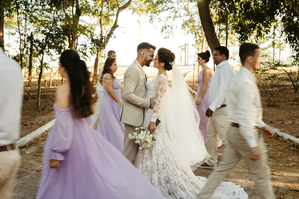 Bride in a white lace wedding gown and veil holding a bouquet, standing with a groom in a light beige suit, surrounded by their wedding party including bridesmaids and best man.